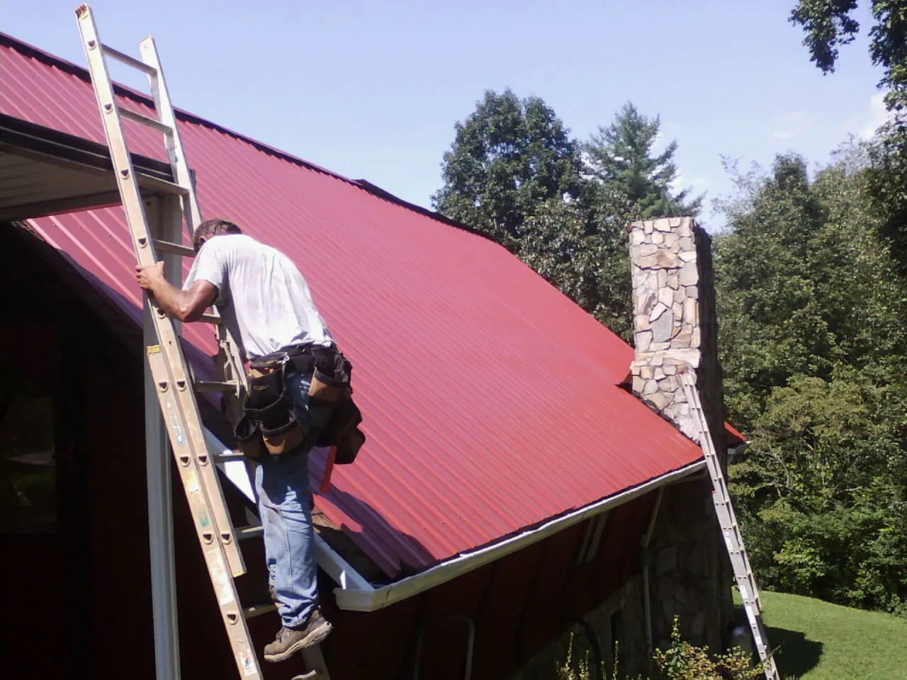 Roofer on a ladder beside a red metal roof and stone chimney — Soffit & Fascia Repair by Roofing Brothers in Gunpowder